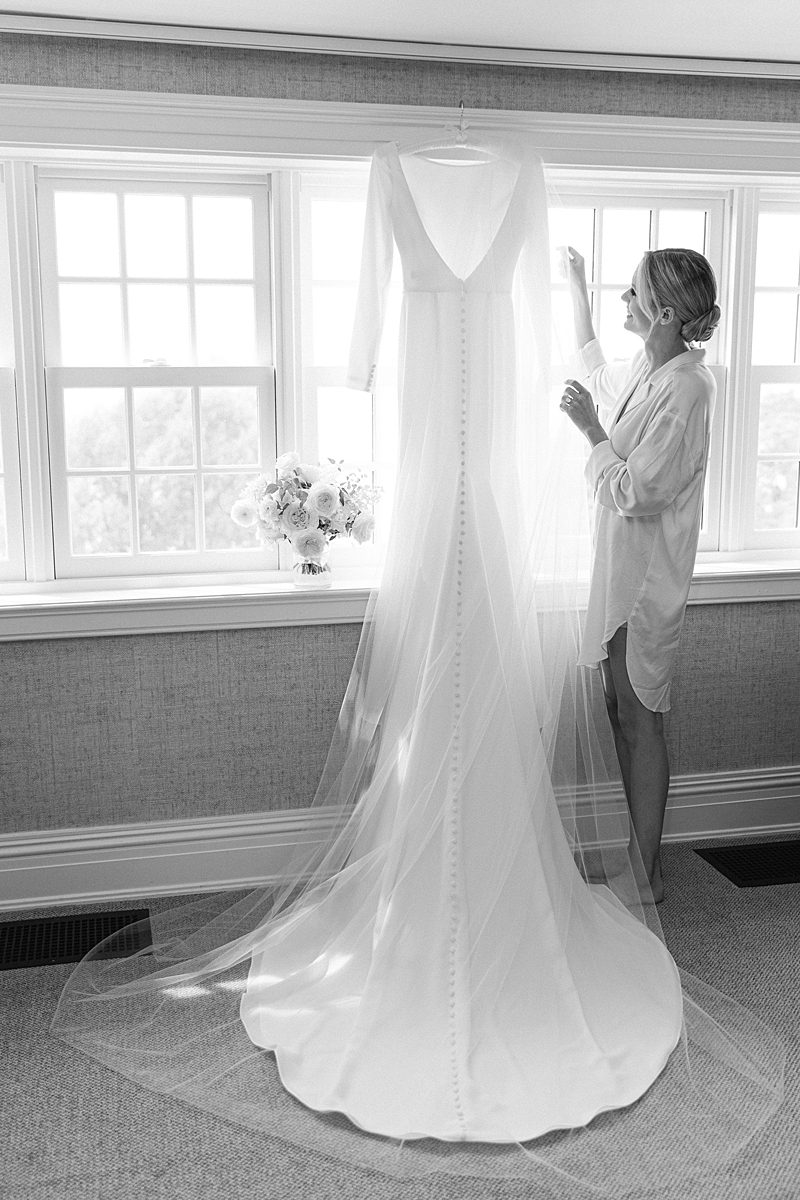 BLACK AND WHITE PHOTO OF BRIDE ADMIRING WEDDING DRESS HANGING UP IN WINDOW